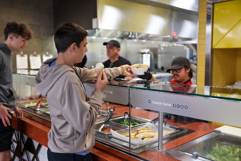 A student takes a pasta bowl from a server while holding an ice cream cone